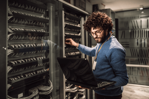 A man with curly hair holding and looking at a laptop in a room with servers