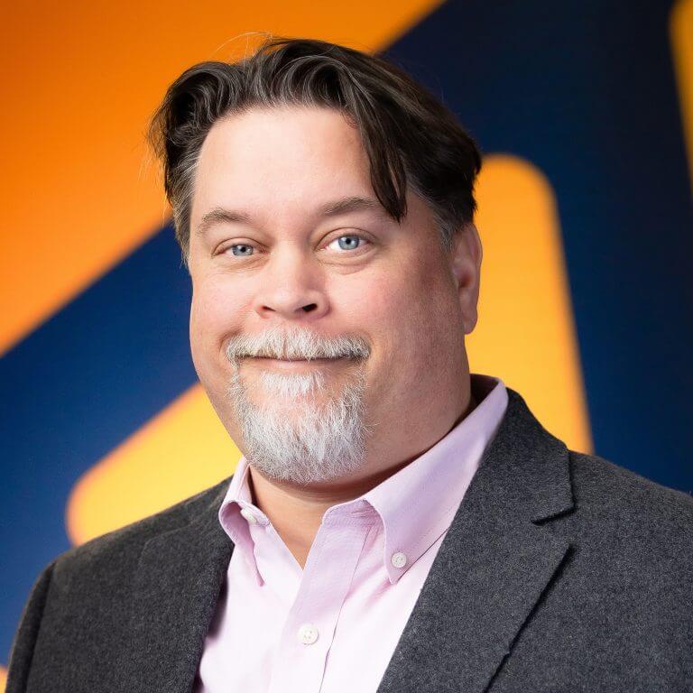 A many in gray suit, pink shirt, in front of blue and orange background.